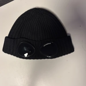 Svart  mössa från C.P. Company - Snygg svart ribbad mössa från C.P. Company med unika glasögondetaljer framtill. Perfekt för att ge din outfit en cool touch. Tillverkad i ett bekvämt material som håller dig varm. Tveka ej på att höra av dig vid minsta intresse eller fråga!🤗