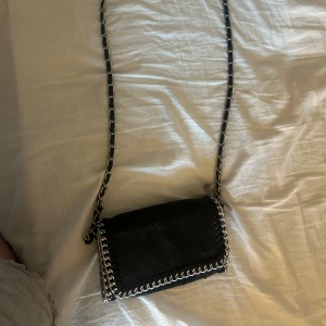 Svart axelväska med kedjedetaljer från Tiamo - Säljer en svart axelväska från Tiamo med snygga silverfärgade kedjedetaljer runt kanterna och på axelremmen. Väskan är i skinnimitation och har magnetlås. Perfekt för att lyfta vilken outfit som helst.