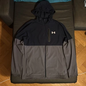 Under Armour tvåfärgad vindjacka med huva - Snygg vindjacka från Under Armour i svart och grått. Jackan har huva, dragkedja framtill och två sidofickor med dragkedja. Perfekt för blåsiga dagar och sportiga outfits. Under Armour-logga på bröstet.