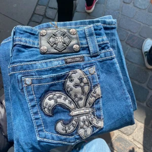 Blå jeansbyxor från Miss Me med broderad ficka - Säljer ett par blå jeansbyxor från Miss Me med en stor, broderad fleur-de-lis på bakfickan och dekorativa detaljer i silver. Byxorna har bootcut-modell och markerade sömmar. Perfekta för dig som vill sticka ut med snygga detaljer. 