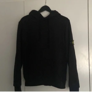 Svart hoodie från Stone Island - Säljer en svart hoodie från Stone Island med deras ikoniska märke på ärmen. Tröjan har en klassisk design med huva och långa ärmar. Perfekt för en stilren och avslappnad look.
