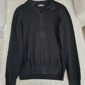 Svart tröja med dragkedja från Dressmann - Säljer en stilren svart tröja från Dressmann med dragkedja och ståkrage. Perfekt för en avslappnad men ändå snygg look. Tröjan har långa ärmar och knappar vid kragen för extra detalj.