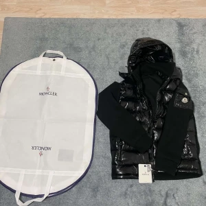 Moncler Shiny Cardigan - Säljer en svart glansig dunväst från Moncler med stickade svarta ärmar och huva. Västen har dragkedja framtill, Moncler-logga på bröstet och medföljande skyddspåse. Perfekt för lager-på-lager och streetstyle.
