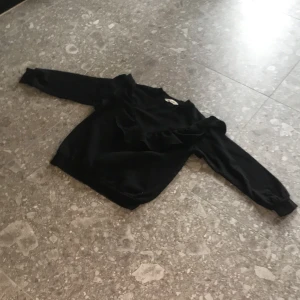 Svart tröja med volang från H&M - Säljer en svart långärmad tröja från H&M i storlek 134/140. Tröjan har en snygg volangdetalj framtill som ger en cool touch. Perfekt för dig som gillar stilrena plagg med en twist.