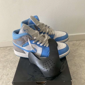 Nike Air Jordans Mid blå och grå - Säljer ett par Nike Air Jordans Mid i blått, grått och vitt. Skorna har klassisk snörning, rund tå och är tillverkade i skinn och syntet. Perfekta för dig som gillar streetwear och vill sticka ut med snygga färgblock.