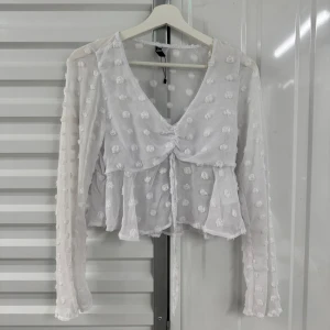 Vit blus med volang och knyt - Säljer en söt vit blus med volang och knyt framtill. Blusen har långärmade transparenta ärmar och är dekorerad med små broderade prickar. Perfekt för dig som gillar romantisk och trendig stil.