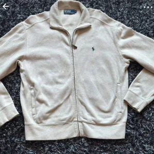 Beige ziptröja från Polo Ralph Lauren - Säljer en beige ziptröja från Polo Ralph Lauren med hög krage och broderad logga på bröstet. Tröjan är tillverkad i 100% bomull och har långa ärmar samt ribbade muddar. Perfekt för en stilren och avslappnad look.