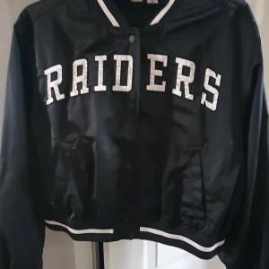 Svart Raiders bomberjacka från H&M x NFL - Säljer en svart bomberjacka från H&M i samarbete med NFL, med stora vita RAIDER-tryck fram och broderad logga på ryggen. Jackan har ribbade muddar, knappar och två fickor framtill. Perfekt för dig som gillar sportig stil.