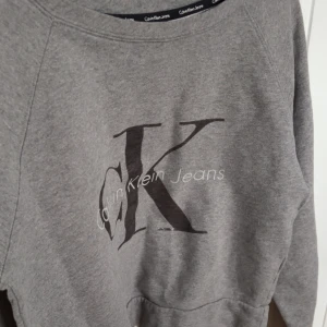 Grå sweatshirt från Calvin Klein Jeans - Grå sweatshirt från Calvin Klein Jeans med stor CK-logga framtill. Tröjan har rund halsringning och långa ärmar. Perfekt för en avslappnad och stilren look.