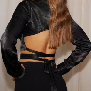Svart croppad blus från Nelly - Snygg svart croppad blus i satin från Nelly med långa ärmar och knytband i ryggen. Oanvänd storlek s