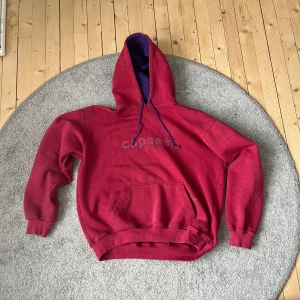 Röd hoodie från Capeesh - Säljer en snygg röd hoodie från Capeesh med broderad text på framsidan. Hoodien har en stor ficka framtill och en justerbar huva med lila snören. Insidan av huvan är lila vilket ger en cool kontrast. Perfekt för en avslappnad stil!