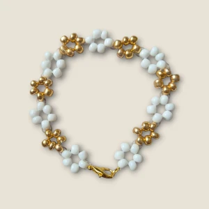 Pärlarmband med blommor i vitt och guld - Superfint armband med små blommor gjorda av vita och guldfärgade pärlor. Armbandet har ett klassiskt lås i guldton och passar perfekt för att ge en söt touch till din outfit.