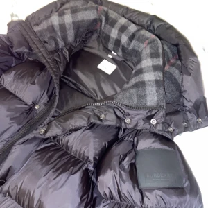 Svart pufferjacka från Burberry - Säljer en snygg svart pufferjacka från Burberry med klassiskt rutigt foder. Jackan har en hög krage och dragkedja framtill. Perfekt för kyliga dagar med stil.sitter S