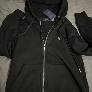 Svart hoodie från Polo Ralph Lauren - Säljer en stilren svart hoodie från Polo Ralph Lauren med dragkedja och det klassiska logotypbroderiet på bröstet. Perfekt för en avslappnad stil. Hoodien har långa ärmar och en mjuk insida för extra komfort.