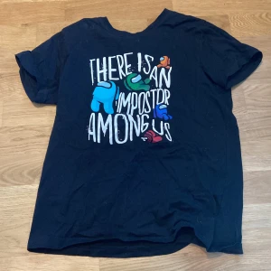 Svart t-shirt med Among Us-tryck - Säljer en svart t-shirt med ett coolt Among Us-tryck i flera färger. Texten 'There is an impostor among us' pryder framsidan tillsammans med karaktärer från spelet. Perfekt för fans av spelet!