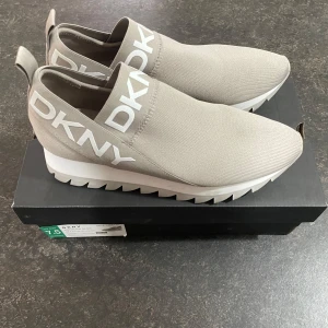 Beige sneakers från DKNY - Snygga beige sneakers från DKNY med vit sula och tydlig logga på sidan. Skorna har en elastisk passform och är perfekta för en stilren look. De har en rund tå och är tillverkade i syntetmaterial.