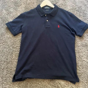 Mörkblå pikétröja från Ralph Lauren - Snygg mörkblå pikétröja från Ralph Lauren med klassisk krage och två knappar. Den har en liten röd broderad logga på bröstet. Perfekt för en stilren look!          Nästan nyskick andvänt 2 gånger men var lite för stor på mig.             Storlek s och passar ca 180 mer eller mindre Mvh 