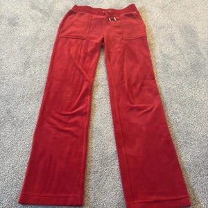 Röda Juicy couture byxor med snörning - Snygga röda byxor med snörning i midjan och praktiska fickor framtill. Byxorna har en rak passform och är tillverkade i ett mjukt material som ger en bekväm känsla. Perfekta för en avslappnad stil. Dem har två fläckar men syns inte så tydligt. Använd fåtal gånger. 