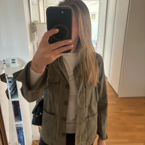 Kahki jacka från Zara - Säljer en kahki jacka från Zara med broderade detaljer på ryggen. Jackan har långa ärmar och knappar framtill. Perfekt för en avslappnad stil med en unik touch.