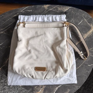 Vit axelväska från Marc by Marc Jacobs - Säljer en stilren vit axelväska från Marc by Marc Jacobs i mjukt skinn. Väskan har justerbar axelrem, guldfärgade metalldetaljer och en diskret logoplatta framtill. Perfekt för dig som vill ha en klassisk och rymlig accessoar.