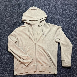 Grå hoodie med dragkedja från Polo Ralph Lauren - Säljer en stilren grå hoodie från Polo Ralph Lauren med dragkedja och huva. Tröjan har två fickor framtill och en broderad logga på bröstet. Perfekt för en avslappnad look. ❗️endast tröjan går med i köpet 