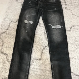 Svarta slitna jeans från Dsquared2 - Säljer ett par svarta jeans från Dsquared2 med slitningar och hål på benen. Jeansen har fem fickor, dragkedja och knapp, samt röda detaljer på insidan. Snygg tvätt och trendig look.