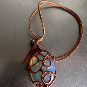 Halsband med opal och brun läderrem - Unikt halsband med en stor opal-liknande sten infattad i konstfullt lindad koppartråd. Hängs i en brun läderrem med dekorativ detalj vid knuten. Perfekt för dig som gillar bohemisk stil och naturinspirerade smycken.