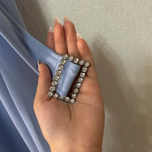 Ljusblå jumpsuit med bälte - Elegant ljusblå jumpsuit med axelband och ett matchande bälte med glittrande spänne. Jumpsuiten har en dragkedja i ryggen för enkel påklädning. Perfekt för speciella tillfällen.