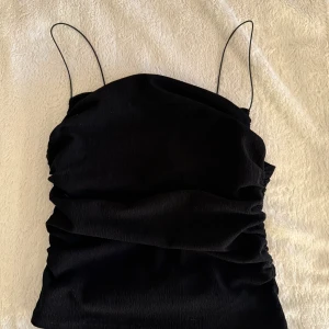 Svart croppad linnetopp med tunna axelband - Säljer en svart croppad linnetopp med tunna axelband och draperad effekt. Perfekt för att styla med jeans eller kjol. Enkel och stilren design som passar till många tillfällen.