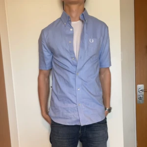 Ljusblå kortärmad skjorta från Fred Perry - Säljer en ljusblå skjorta från Fred Perry med korta ärmar och klassisk button-down krage. Skjortan har vita knappar och en broderad Fred Perry-logga på bröstfickan. Perfekt för en clean och avslappnad look. Nyskick endast använt 1 gång. Killen är 184.
