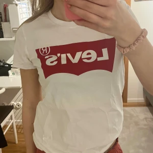 Vit t-shirt med röd Levi's-logga - Säljer en klassisk vit t-shirt från Levi's med den ikoniska röda loggan tryckt på bröstet. T-shirten har rund halsringning och korta ärmar. Perfekt basplagg för garderoben!