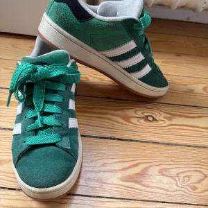Adidas Campus gröna sneakers - Säljer ett par gröna Adidas Campus sneakers med vita ränder och detaljer. Skorna har ovandel i mocka, klassisk Adidas-logga på hälen och gröna skosnören. Snygg retrostil som passar till många outfits.