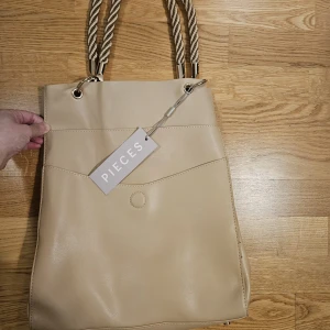 Beige axelväska från Pieces med repdetaljer - Snygg beige axelväska från Pieces med grova rep som axelband och guldfärgade metalldetaljer. Väskan är rymlig och har en enkel, stilren design med magnetknäppning. Perfekt för dig som vill ha en stilren accessoar till vardagslooken.