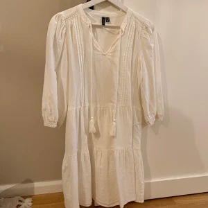 Vit klänning med knyt från Vero Moda - Säljer en luftig vit klänning från Vero Moda med trekvartsärm och knytband med tofsar vid halsen. Klänningen har volangdetaljer och lätt v-ringning, perfekt för varma dagar.