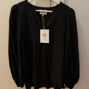 Svart blus - Säljer en svart blus från nümph med v-ringning och knappar framtill. Blusen har långa, lätt puffade ärmar och är tillverkad i ett mjukt, lätt material som ger ett snyggt fall. Perfekt för dig som gillar stilrena plagg med en elegant touch.