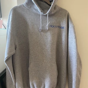 Grå hoodie Harry's House  - Grå hoodie från Harry Styles med blått tryck fram och stort blått motiv på ryggen. 