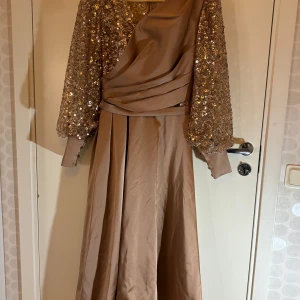 Champagnefärgad långklänning med paljetter - Säljer en elegant långklänning i champagnefärg med glittriga paljetter på överdelen och ärmarna. Klänningen har en axelbandslös design med draperad detalj framtill och långa ärmar. Perfekt för bal eller fest.
