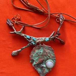 Handgjort halsband med blad och sten - Unikt halsband med ett konstnärligt hänge format som en gren och ett blad i grönt och kopparfärgat. Hänget har en rund vit sten och detaljer i turkos. Långt brunt läderband med kopparfärgade metalldelar.