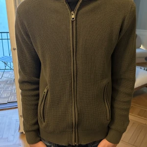 Indicode cardigan - Säljer en olivgrön indicode jeans kofta/cardigan. Koftan har ribbade muddar och en huva i ett annat material. Huvan är även avtagbar om man önskar det. Perfekt nu inför våren. Snygga till jeans! Modellen är 193cm 82kg. Hör av er vid eventuella funderingar!