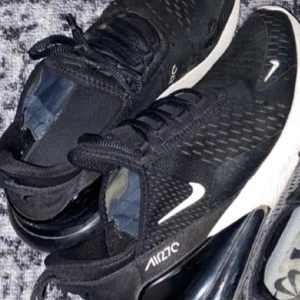 Svarta Nike Air Max 270 sneakers - Säljer ett par svarta Nike Air Max 270 sneakers med vit sula och tydlig Nike-logga på sidan. Skorna har snörning och en luftig mesh-överdel. Perfekta för en sportig och stilren look.