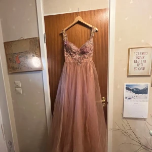 Rosa balklänning med broderier från Gino Cerruti - Säljer en fantastisk långklänning från Gino Cerruti i en dammig rosa nyans. Klänningen har smala axelband, v-ringad urringning och är dekorerad med broderade blommor och pärlor upptill. Kjolen är i flera lager tyll för en drömmig look.