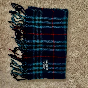 Rutig halsduk från Burberry - Säljer en klassisk rutig halsduk från Burberry i 100% kashmir. Halsduken har blå, svarta och röda rutor samt fransar i ändarna. Perfekt accessoar för att lyfta din outfit.