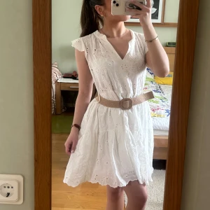 Vit kortklänning med spets och bälte - Söt vit klänning med broderade blommor, v-ringning och volangärmar. Perfekt till midsommar! Köpt i Spanien 