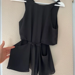 Svart ärmlös blus  med knytband - Snygg svart ärmlös blus med knytband i midjan. Perfekt för en stilren look. Blusen har en enkel design med rund hals och är tillverkad i ett lätt material.