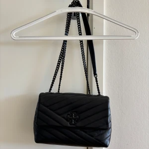 Tory Burch väska  - Snygg svart axelväska från Tory Burch i quiltat skinn. Väskan har en elegant kedjerem och ett stilrent märke på framsidan. Perfekt för att ge din outfit en lyxig touch.