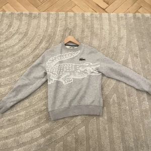 Grå sweatshirt från Lacoste - Säljer en grå sweatshirt från Lacoste med en stor vit krokodil på framsidan. Tröjan har en loose fit och långa ärmar, perfekt för en avslappnad stil. Klassisk och stilren design med ribbade muddar vid ärmslut och nederkant. Storleken på tröjan är xs men sitter ganska löst så passar även S och M .Pris kan diskuteras vid snabb affär!!!