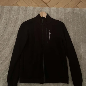 Svart tröja  från Sail Racing - Snygg svart jacka från Sail Racing med dragkedja framtill. Jackan har en stilren design med långärmade ärmar och en hög krage. Perfekt för en sportig look. 