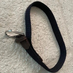 Blått bälte från John Henric - Snyggt blått bälte från John Henric med en stilren design. Bältet har en silverfärgad spänne och bruna läderdetaljer. Perfekt för att ge en elegant touch till din outfit.