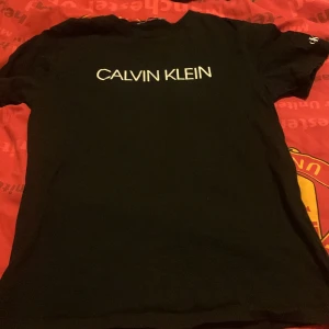 Svart t-shirt från Calvin Klein - Snygg svart t-shirt från Calvin Klein med deras ikoniska logga tryckt på framsidan. Perfekt för en stilren och avslappnad look. Tillverkad i mjukt material för extra komfort.