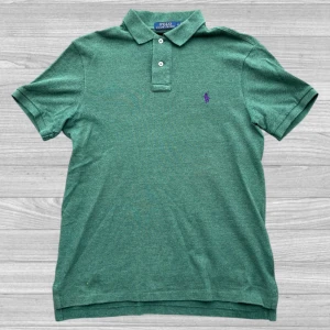 Ralph Lauren piké - Säljer en klassisk grön pikétröja från Ralph Lauren med lila broderad logga på bröstet. Tröjan har korta ärmar och knappar vid kragen. Perfekt för en stilren look!